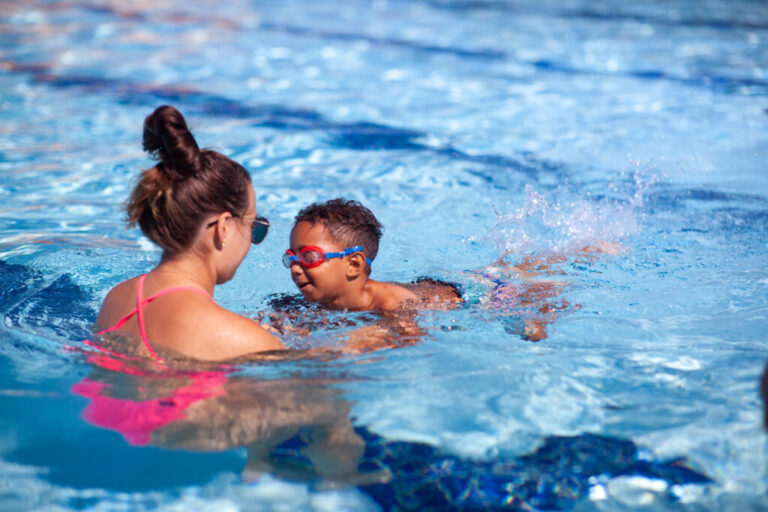 A swim instructor teaches a 3-year-old boy how to swim in an outdoor swimming pool.