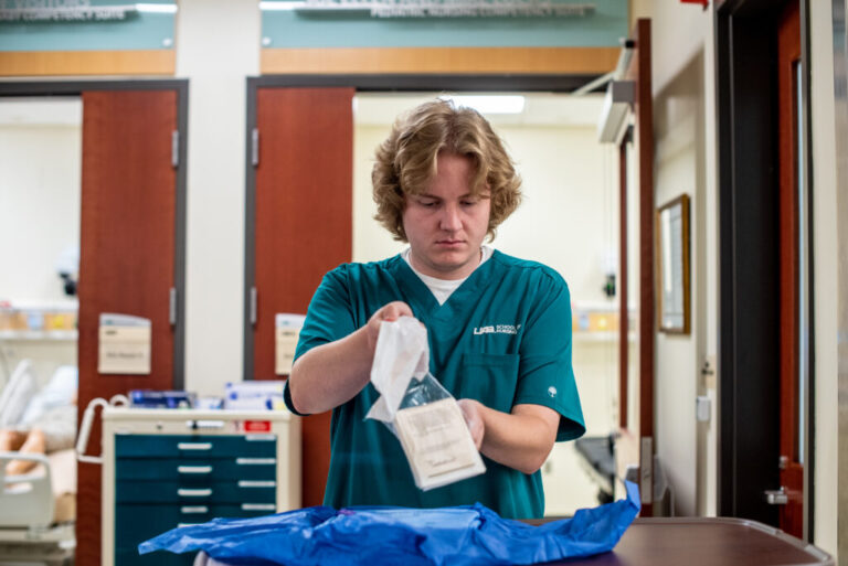 Nursing student Ethan Stinson prepares personal protective equipment during a class exercise. He has blonde curly hair and is wearing teal scrubs.