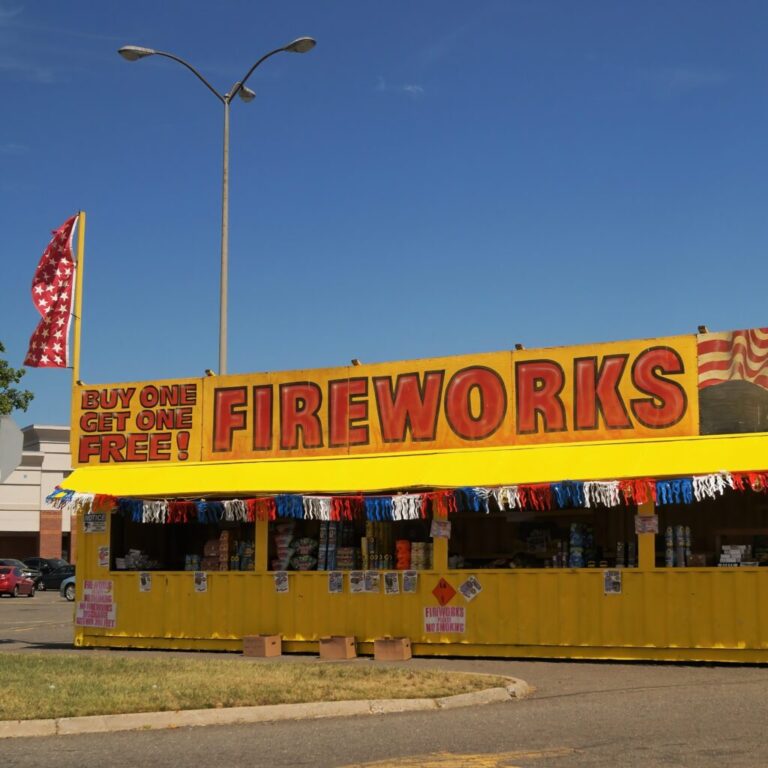 A bright yellow fireworks stand is under a clear blue sky with a large sign at the top of the stand the that reads