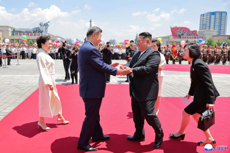 Two leaders wearing suits shake hands outdoors on a red carpet.