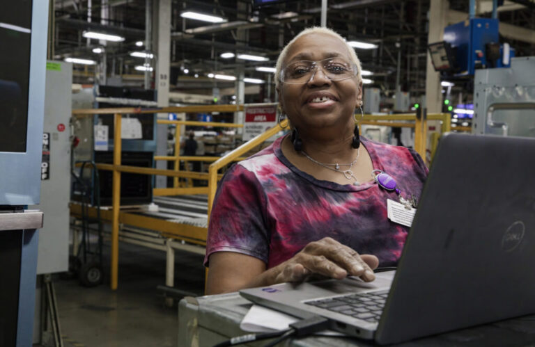 Ruth Ransom, 68, is a woman with short white hair. She is wearing safety glasses and standing behind a laptop on the factory floor.