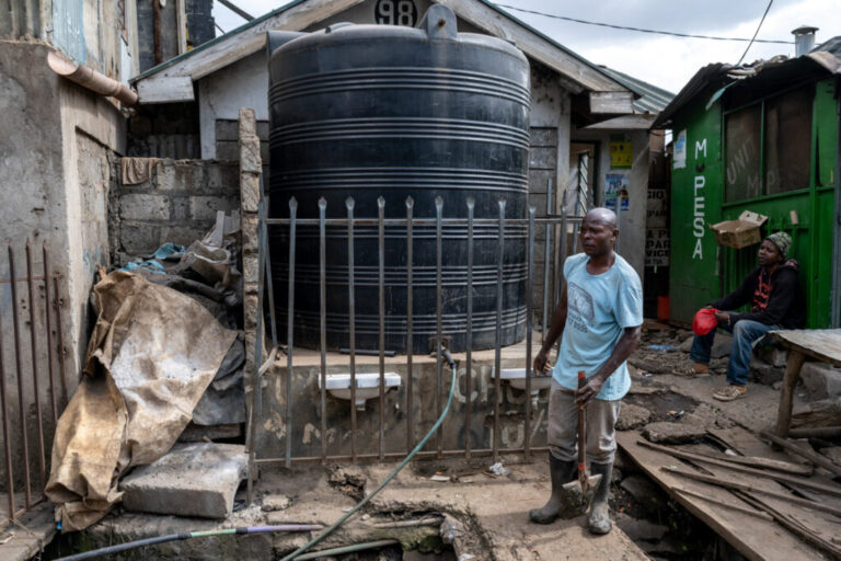 A man wearing rubber boots and holding a shovel stands in front of a water tank with a hose on it.