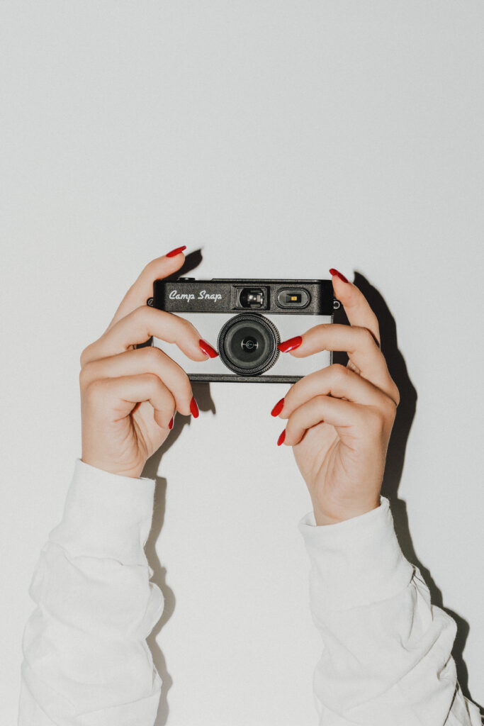 This photo shows a beige and black Camp Snap camera that is being held by a woman's hands, which have long red-pained fingernails.