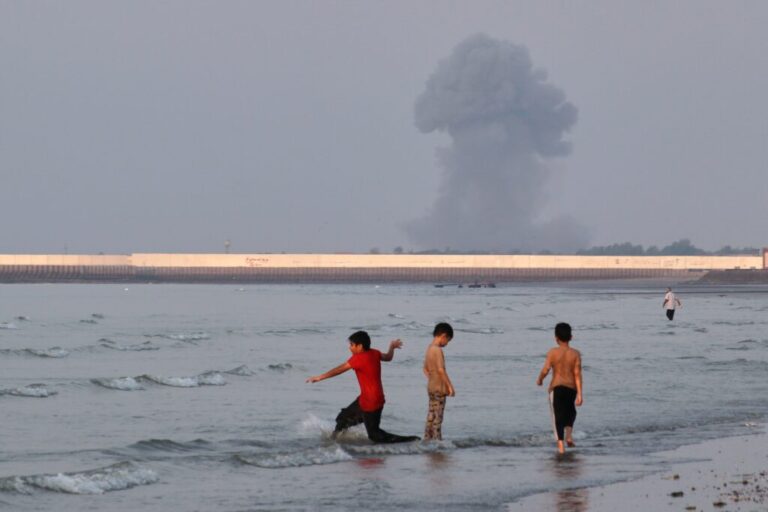 Three boys play in the shallow waters of the Strait of Hormuz, as a plume of smoke rises from an explosion in the background, off Bandar Abbas, Iran, Monday.