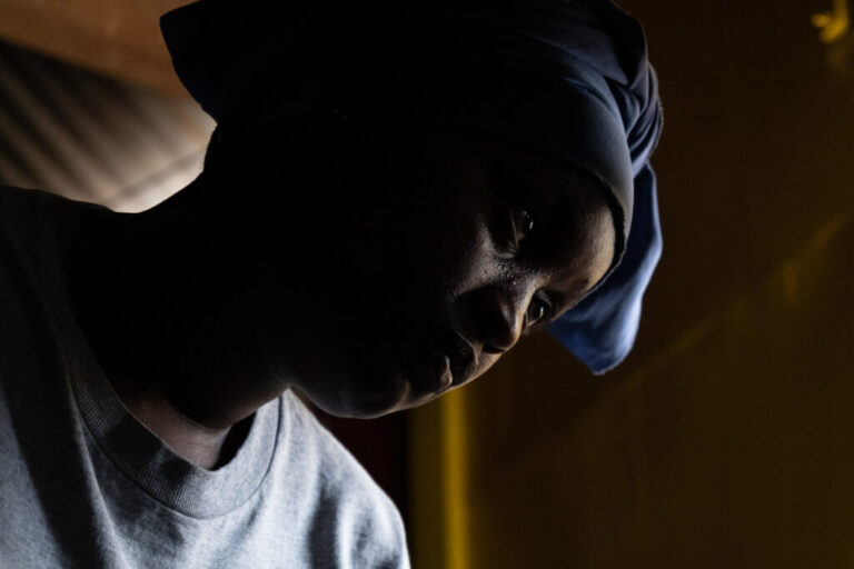 A close-up shows a woman's face and shoulders. She's wearing a gray T-shirt and a blue head covering.