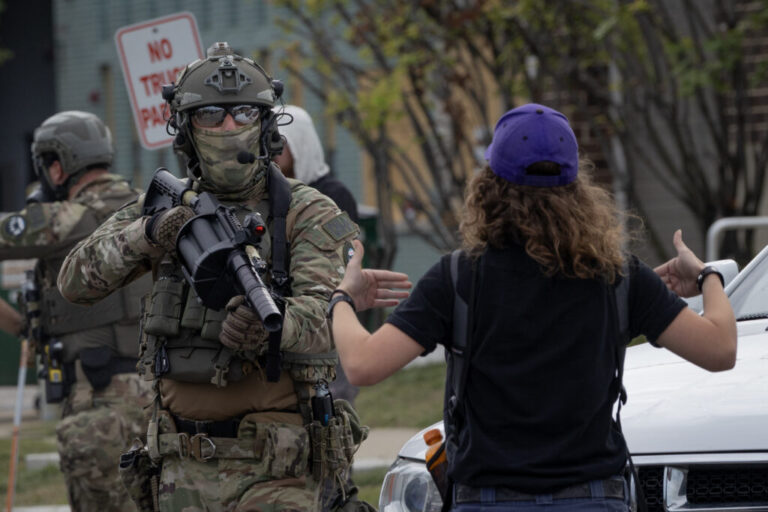 A federal law enforcement agent dressed in camouflage clothing confronts a demonstrator wearing a blue baseball cap with her back to the camera.