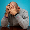 Photograph of a man sitting with his elbows on a wooden table and face palming due to distress and overwhelm. He holds his glasses in his hands. He sits in front of a blue backdrop.