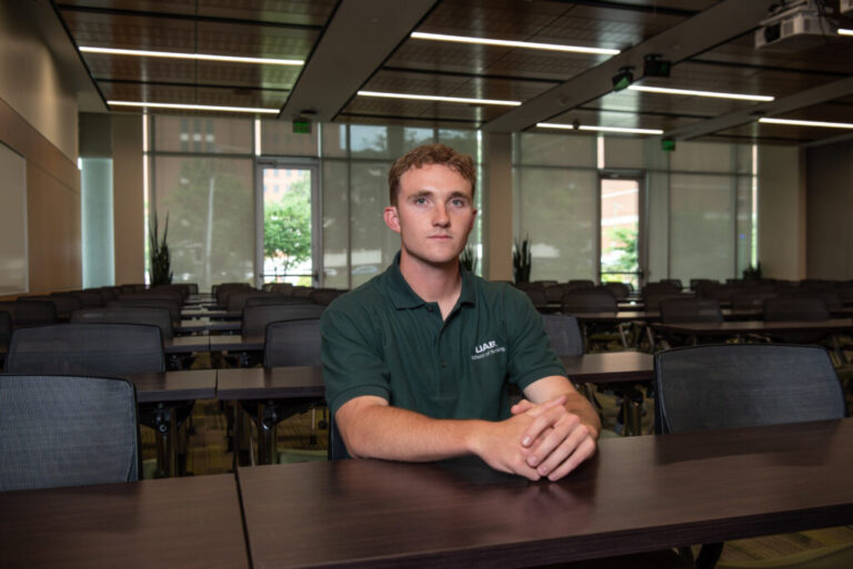 Nursing student Connor Aderholt has short curly hair and is wearing a UAB polo shirt. He is sitting at a table in a classroom.