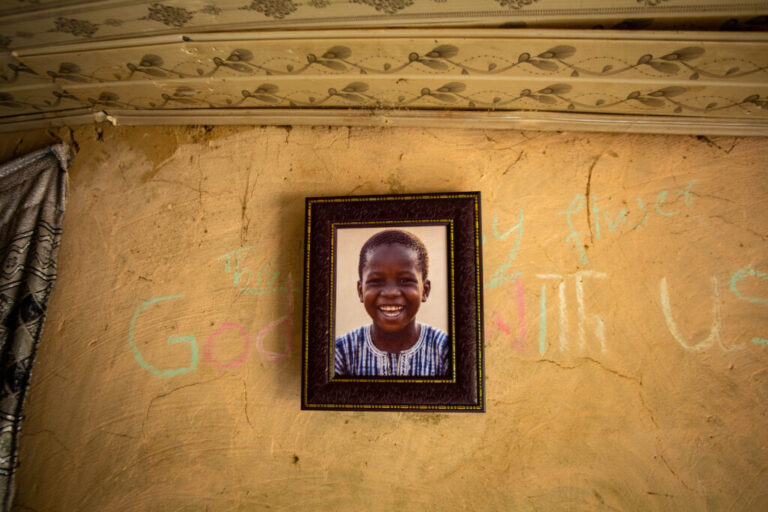 A framed photo of Ibrahim Garba hangs on a wall inside his family’s home. In the photo, he has a wide smile. The words “God is with us,