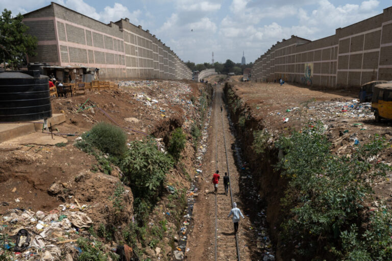 People walk along train tracks running between two high walls. There's trash on the ground all around.