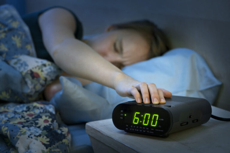 A young woman who's lying in bed under a blanket reaches out her arm to press the snooze button on a digital clock radio that's on a nightstand that's next to the bed.