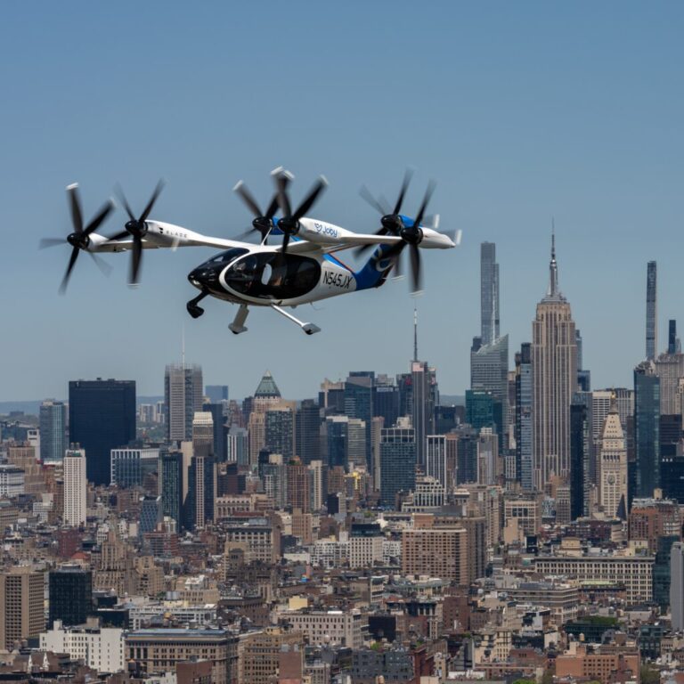 Buildings in the background against a blue sky, where a black and white air-taxi flies over the New York City skyline.