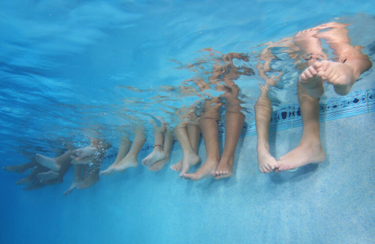 This photo, taken underwater, shows the lower legs of children who are sitting in a row along the edge of a swimming pool. Their legs, from the knees on down, are dangling in the pool's water.