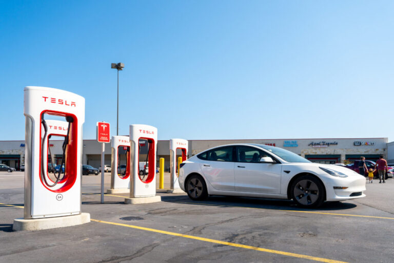 Tesla Superchargers stand in a parking lot in Austin, Texas, on September 16. Four of the five white and red Superchargers in the photo are unoccupied, and a white Tesla sedan is parked in front of one. A strip mall stretches across the background.