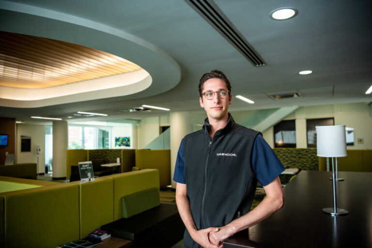 Graduate nursing student Chris Jones is wearing a black vest over his blue scrubs. He has short brown hair and is wearing glasses while standing in the school's lounge area.