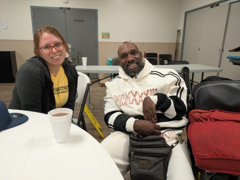 Dr. Elyse Stevens wears glasses, a yellow t-shirt and a sweater. Ronald Major wears a stylish white track suit.