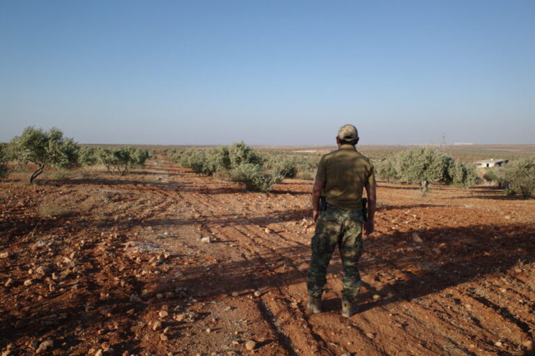 With his back to the camera, a senior Uyghur militant in camouflage-print pants stands facing an olive grove in northern Syria.