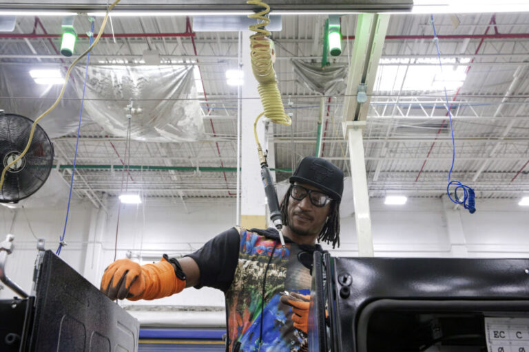 Kwame Crockett is wearing orange safety gloves, safety glasses, a black baseball cap and a colorful shirt as he assembles parts of an oven.