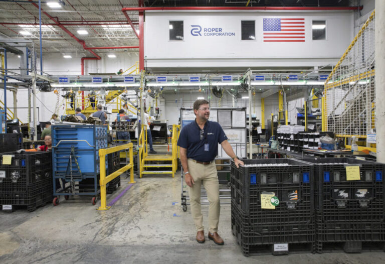 Tony Gabbert is standing on the factory floor, wearing safety glasses, a navy polo shirt and khaki pants. Workers are going about their jobs behind him.