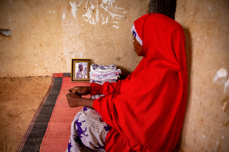 Fatima Ibrahim sits on a mat on the floor in her home. At the other end of the mat, propped up against a wall, is a framed portrait of her deceased son, Abdullahi Ibrahim, a young boy wearing a brown outfit.