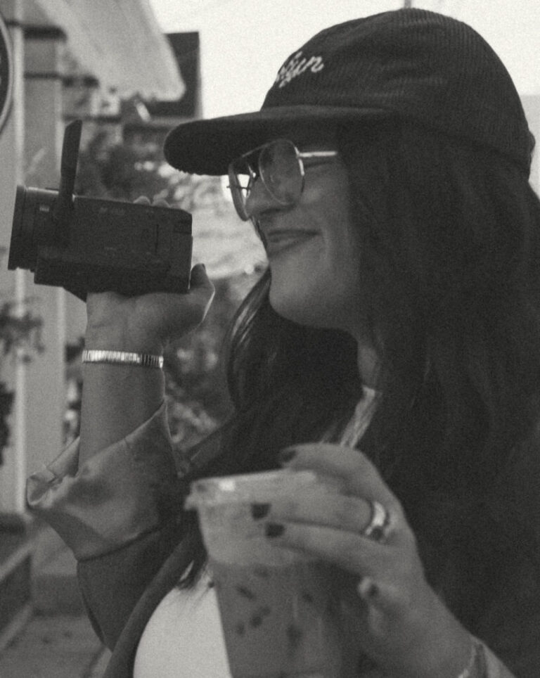In this black-and-white photo, Christina Berkett is holding a camcorder in one hand and a drink in a to-go cup in the other. She's wearing a baseball cap and glasses, and she's smiling.
