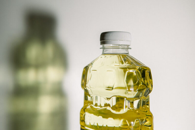 A close-up photograph of canola oil in a plastic bottle. The shadow of the bottle is cast to the left of the bottle on the white wall behind the bottle of seed oil.