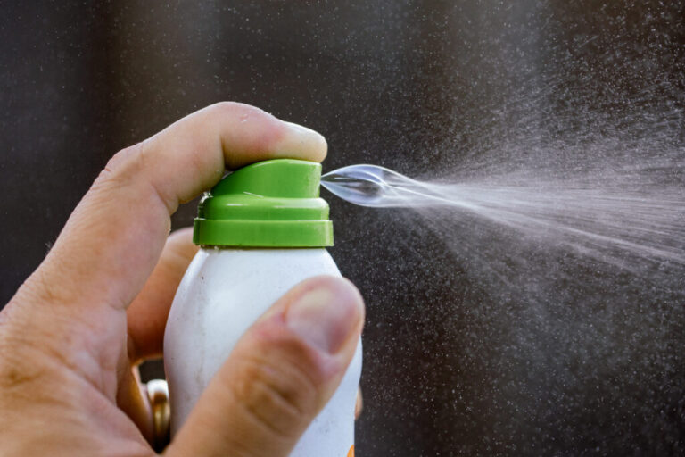 Close-up photograph showing a hand pressing down the button on a spray can of mosquito repellent. The spray catches the light as it's misted out of the can.