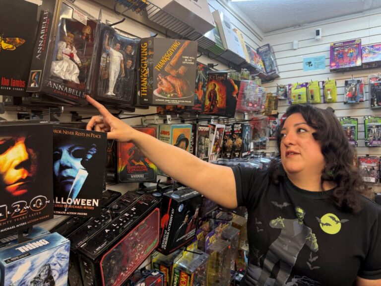 A woman points at a box containing an Annabelle doll displayed on the pegboard wall of a toy store.