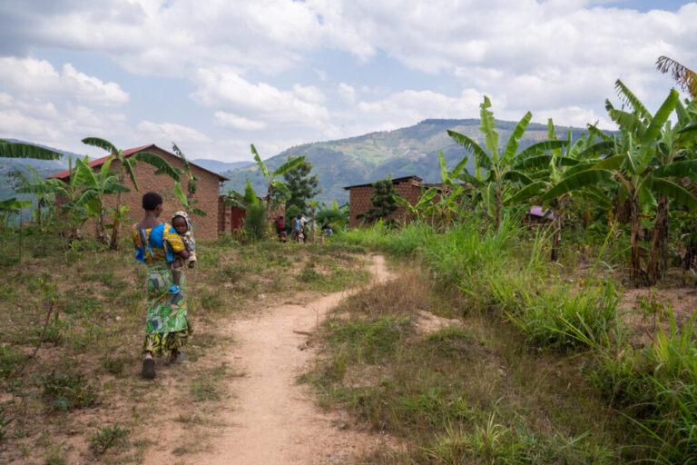 A woman holding a baby walks along a narrow dirt path that leads to Mukanoheli's village. There are several small homes with palm trees growing nearby and a mountain in the distance.