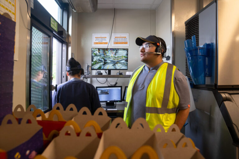 Ebert Velasquez Ramos (right) works at the drive-through at a McDonald's in Morningside, Md. The recent high school graduate is wearing a yellow safety vest over his gray polo uniform. He is also wearing a visor and headset to communicate with customers. Another employee has her back to the camera and there are several empty Happy Meal boxes in front of them.