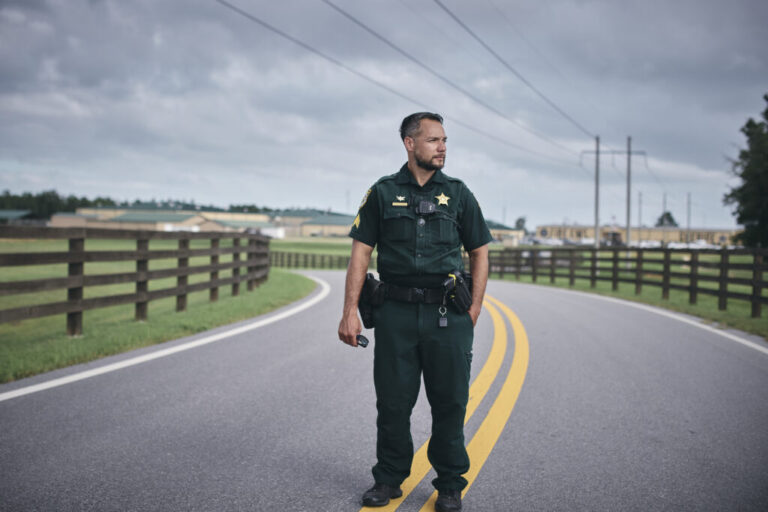 Walton County Sheriff's Office Sgt. Oscar D’Lassalas, wearing his uniform, stands in the middle of a road, on its two yellow parallel dividing lines. A wooden fence is on each side of the road.