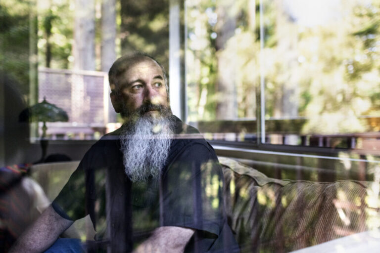 In this photo, David Davis is seated on a brown sofa with his head turned toward the right side of the frame. He is wearing a dark T-shirt and has a long beard.
