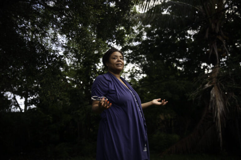 Wearing a dark blue dress, Farah Larrieux stands in her backyard in South Florida. Multiple trees stand in the background.