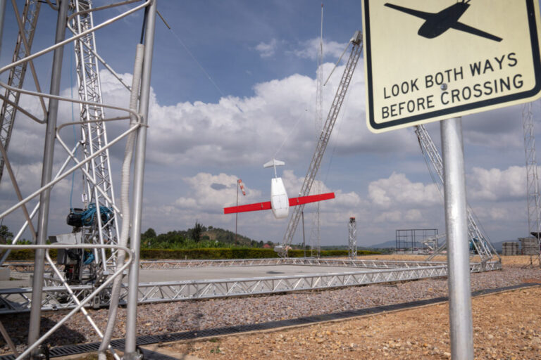 A drone, just returned from a delivery, swings in the landing mechanism in Muhanga, Rwanda. The drone is white with red wings. There is a sign that reads