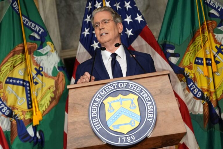 Treasury Secretary Scott Bessent stands behind a lectern equipped with a microphone as he speaks at a news conference at the Treasury Department on Monday. The department's seal is on the front of the lectern, and three flags are behind Bessent: a U.S. flag flanked on each side by a Treasury Department flag.