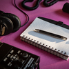 A blank notebook with a pen, a recorder, headphones and a mic are laid out on a bright fuchsia background.