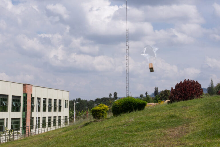 Zipline drones drop packages from the sky which are filled with medicines at Kabgayi Hospital in Muhanga. A cardboard box with a transparent parachute is seen floating towards a grassy hill next to a hospital building.