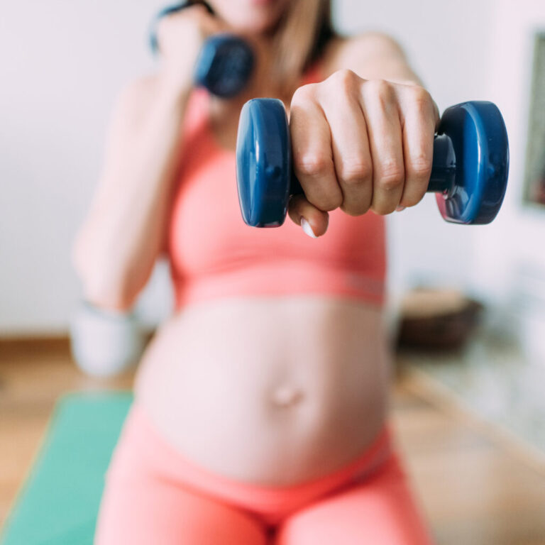 A photograph of an unrecognizable pregnant woman exercising with dumbbells in her living room at home.