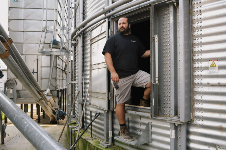 Ian Stamy stands in the doorway of one of his grain silos on August 12. The silo is made from silver corrugated metal. Stamy is wearing boots, light-colored shorts and a dark T-shirt.