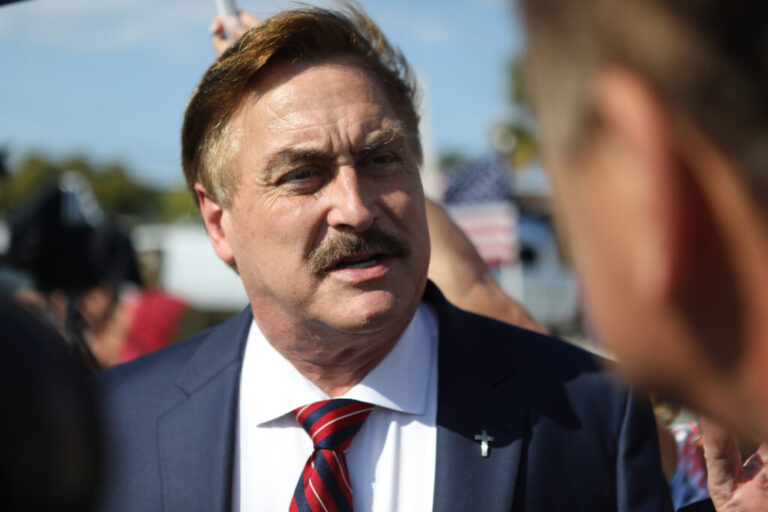 MyPillow CEO Mike Lindell arrives at a gathering of Donald Trump supporters in Palm Beach, Florida, on April 4, 2023. Photographed from about the chest up, he's wearing a blue suit jacket, a white shirt and a red and blue tie.