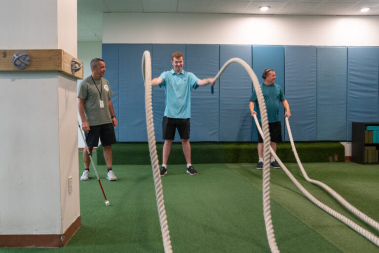 At a gym, Cody holds a thick rope in each hand and moves them in a wave pattern. On the left side of the frame is Next Steps health and wellness coordinator Brian Prendergast. On the right side of the frame is another Next Steps client, who is also holding two ropes.