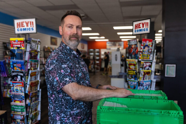 Marc Bowker, owner of Alter Ego Comics in Lima, Ohio, thumbs through a green bin of comic books and either side of him are two shelves with signs that read