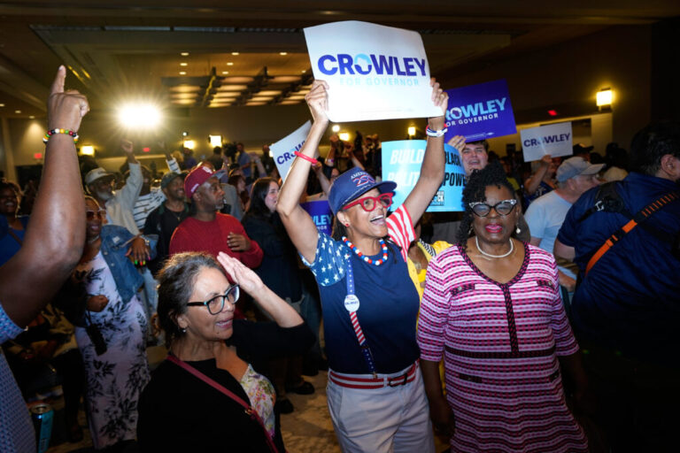 A group of people in a large room cheer and some hold signs above their heads saying,
