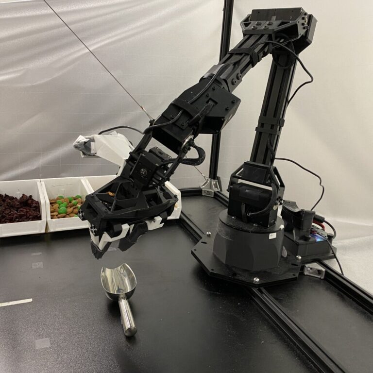 A robotic arm hovers over a metal scoop, placed in front of a series of trays with various snacks.
