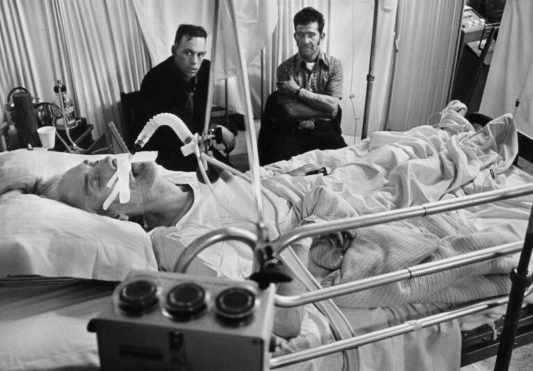 In this 1976 black-and-white photo, coal miner Jess Bishop lies in a hospital bed, with a tube in his throat, as his two adult sons sit on chairs beside his bed.