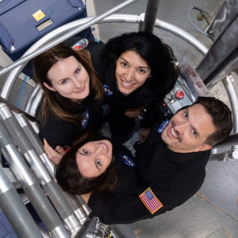 Four people smile up at the camera. They are wearing dark uniforms and one person has a U.S. flag patched onto the sleeve. They are close together, in a small, metallic, well-lit ladder well.