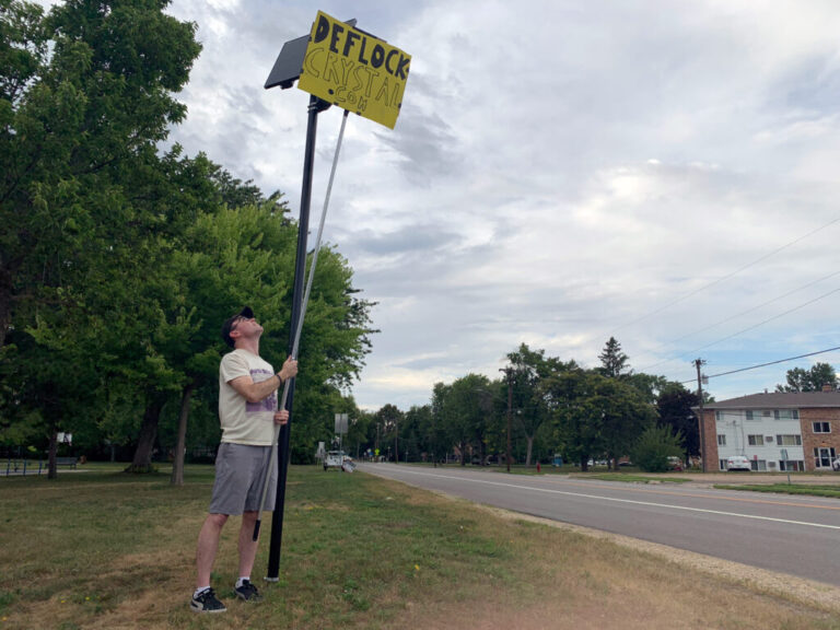 A man uses a long metal pole with a bright yellow sign taped to the end to block the view of a camera mounted on a pole next to a road. The sign says