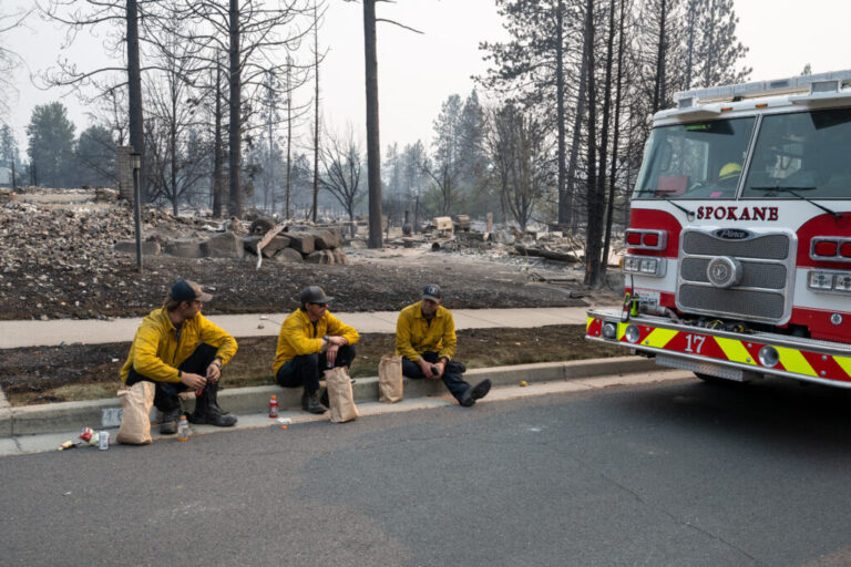 Three members of the Spokane Fire Department sit on a curb in front of burned out residential homes in the Balboa neighborhood of Spokane, Wash., on August 2, 2026.