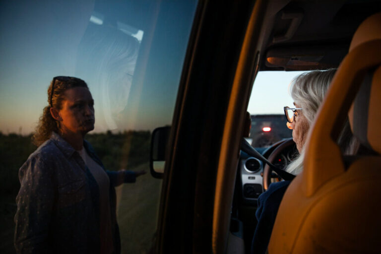 Robyn Smith sits behind the steering wheel of a vehicle as she speaks with Heather Marsh, who is standing outside the vehicle, on the left side of the frame.