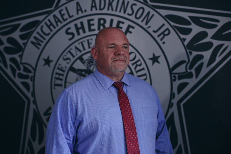 Sheriff Michael Adkinson stands in his Walton County, Florida, office. Photographed from about the waist up, he's wearing a light blue, long-sleeved shirt and a red patterned tie. Behind him on a wall is a large image of a sheriff's star with his name on it.